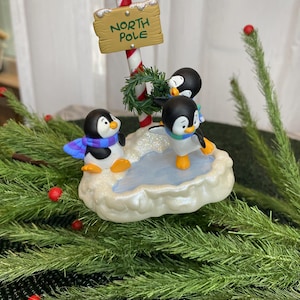 Peut inclure: Décoration de Noël festive avec trois figurines de pingouins sur une base enneigée. Un panneau « North Pole » est soutenu par un bâton de sucre d'orge. Les pingouins portent des écharpes et ont des pieds oranges.
