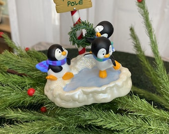 May include: A festive Christmas decoration featuring three penguin figurines on a snowy base. A sign that reads "North Pole" is held up by a candy cane pole. The penguins wear scarves and have orange feet.