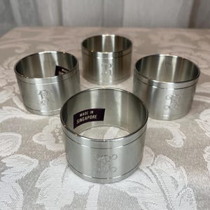 Puede incluir: Cuatro servilleteros plateados con iniciales grabadas. Los anillos son cilíndricos con la parte superior e inferior ligeramente ensanchadas. El texto "MADE IN SINGAPORE" es visible en uno de los anillos. Se encuentran sobre una superficie estampada.
