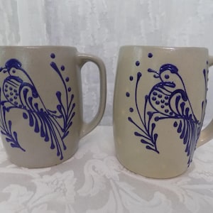 May include: Two beige ceramic mugs featuring a raised blue slip-trailed design of a bird perched on a branch. Each mug has a sturdy handle and a smooth, rounded body. The blue decorative pattern includes dots and stylised foliage surrounding the bird.