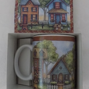 May include: A white ceramic mug featuring a colorful illustration of two houses with trees and a white picket fence, packaged in a matching box that displays the same house scene. The mug has a curved handle and a brown border at the top and bottom.