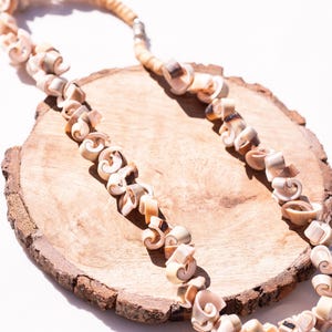 May include: A necklace made of cream-colored, spiral-shaped shells. The shells are strung together, creating a unique and natural design. The necklace rests on a round, wooden surface with a light brown color and a natural edge.