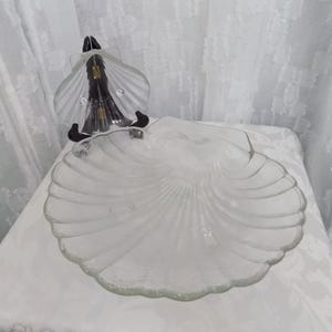 May include: A clear glass serving dish shaped like a large scallop shell with a textured, ribbed surface. The dish is displayed upright on a black stand, showcasing its translucent material and detailed, fan-like radial ridges along the edges.