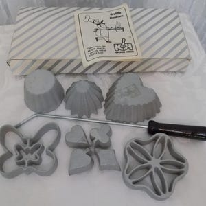 May include: A Kalkus-Hirco waffle baker set featuring a striped box, an instruction manual with a chef illustration, three gray fluted molds, a metal handle with a black grip, and four gray metal cookie cutters in butterfly and card suit shapes.