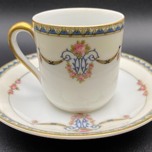 Puede incluir: Un juego de taza y platillo de té de porcelana blanca con borde dorado y diseño floral. La taza tiene un asa dorada y el platillo tiene un borde azul y dorado.