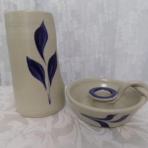 May include: A beige ceramic vase and a matching candle holder, both featuring a hand-painted dark blue leaf motif. The vase is cylindrical with a slightly tapered top, while the candle holder has a circular base and a looped handle.