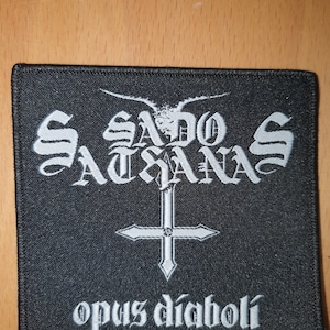 May include: Black square patch featuring white gothic lettering and an inverted white cross. The text reads "Sado Satanas" above the cross and "opus diaboli" below. The patch has a stitched border.
