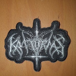 Puede incluir: Parche bordado negro con texto blanco y un diseño de cruz central. El parche presenta la palabra "KATORDA" en una fuente gótica estilizada. El parche es probablemente para una chaqueta o bolso.