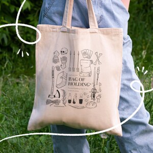 May include: Beige canvas tote bag with black illustrations of fantasy items, including a skull, weapons, and potions. The text "SUPERIOR BAG OF HOLDING" is printed on the front. The bag has two handles and is held by a person.
