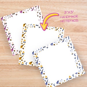 May include: Three 3"x3" handmade notepads with camping-themed borders. The borders feature guitars, campfires, arrows, and crescent moons in purple, yellow, and blue. The notepads are on a light wood surface.