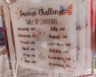 May include: A clear plastic box with a white paper insert that says "Savings Challenge Table of Contents". The paper insert lists each month of the year with a dollar amount next to it. The paper insert has a red, white, and blue American flag design.