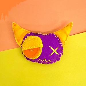 May include: A handmade felt creature with a purple body, yellow horns, and a yellow eye patch. The eye patch has an orange button. The creature has a stitched yellow outline and a stitched yellow "X" on its face. The background is yellow and orange.