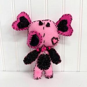 Pode incluir: Boneca de feltro rosa feita à mão com detalhes em preto. A boneca apresenta orelhas pretas em forma de coração, um olho de botão preto e um corpo costurado em preto. Possui um colar de contas e um pequeno pingente de prata. Fundo branco.