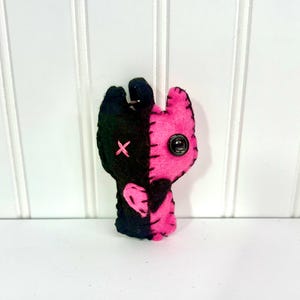 May include: A handmade felt doll with a split design, half black and half pink. The black side features a stitched pink cross, while the pink side has a button eye. The doll is a unique, handcrafted item.