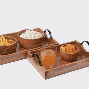 May include: Two wooden serving trays with black metal handles. One tray holds popcorn and french fries in wooden bowls. The other tray holds tortilla chips and a glass of orange juice. The trays are on a white background.
