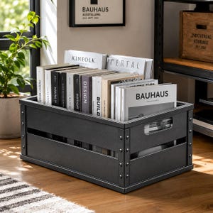 May include: A rectangular dark gray metal storage crate with riveted corner accents and side handle cutouts, filled with a collection of books and magazines featuring titles such as Kinfolk, Design, Build, and Bauhaus on their spines and covers.
