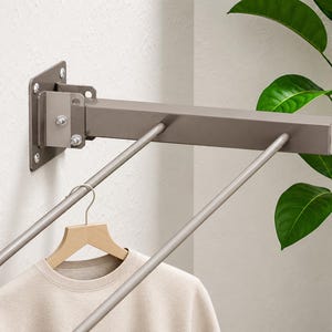 May include: A wall-mounted, taupe-colored metal garment rack featuring a rectangular support arm and two parallel rods extending outward. A light-colored wooden hanger holding a beige crew-neck shirt hangs from one of the metal rods.