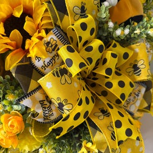 Peut inclure: Gros plan sur une couronne d&eacute;corative avec un grand tournesol jaune, des roses jaunes et du feuillage vert. La couronne est orn&eacute;e de rubans jaunes &agrave; pois noirs et de graphiques d'abeilles. Les rubans portent &eacute;galement les mots "Happy" et "Blessed".