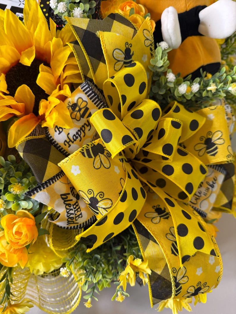 Peut inclure: Gros plan sur une couronne d&eacute;corative avec un grand tournesol jaune, des roses jaunes et du feuillage vert. La couronne est orn&eacute;e de rubans jaunes &agrave; pois noirs et de graphiques d'abeilles. Les rubans portent &eacute;galement les mots "Happy" et "Blessed".