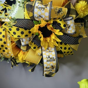 May include: A decorative wreath featuring sunflowers, yellow and black polka dot ribbons, and ribbons with the text "Bee Happy" and "Bee Blessed." The wreath has a bee theme with a cartoon bee illustration.