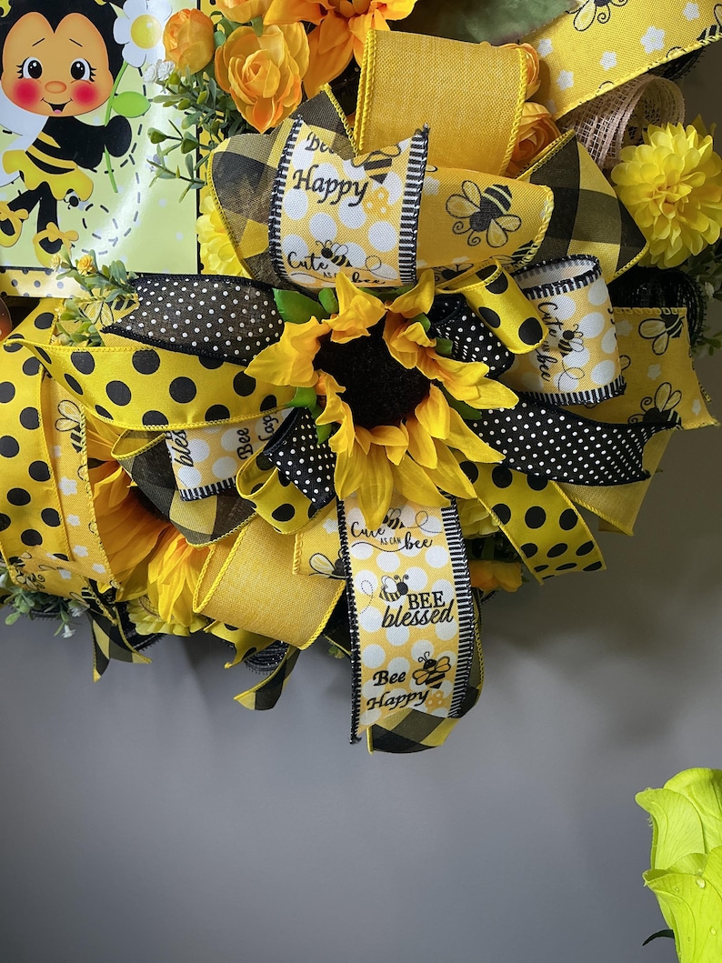 Peut inclure: Gros plan sur une couronne d&eacute;corative sur le th&egrave;me des abeilles, orn&eacute;e de tournesols, de rubans jaunes et noirs &agrave; pois et de rubans avec les textes "Bee Happy" et "Bee Blessed".
