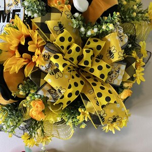 Peut inclure: Une couronne d&eacute;corative avec un tournesol, des n&oelig;uds &agrave; pois jaunes et noirs et une abeille en peluche. La couronne comprend le texte "Bee Blessed" et "Happy". Orn&eacute;e de fleurs jaunes et de verdure.