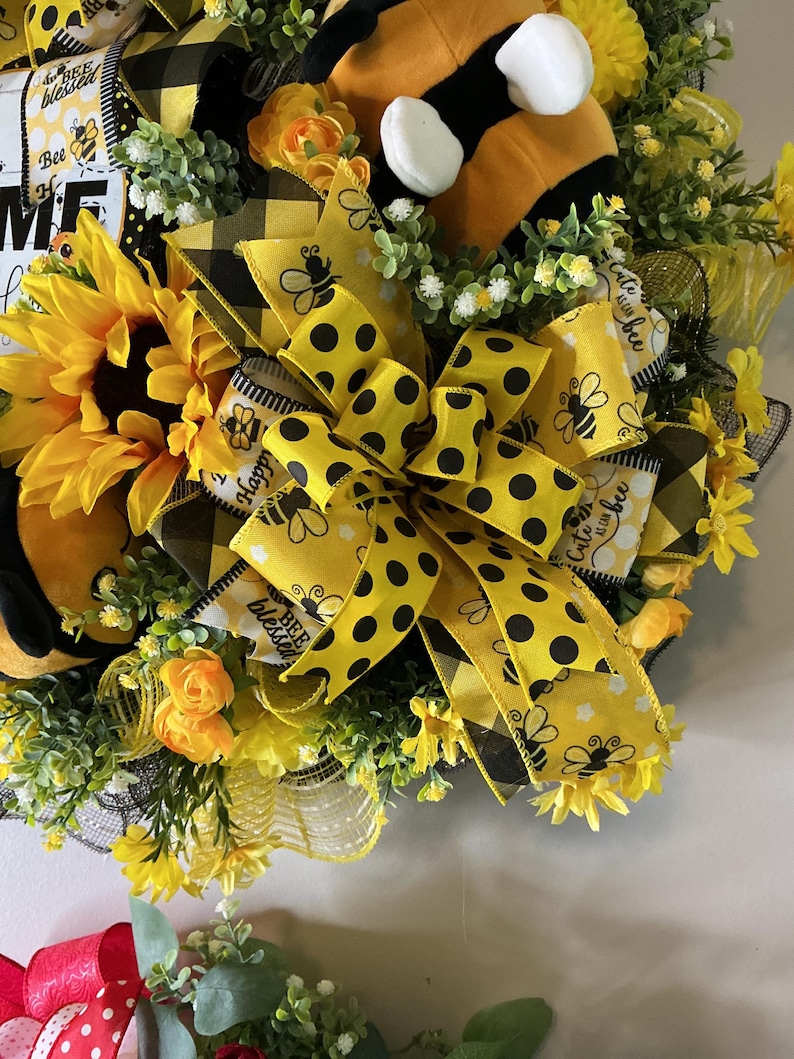 Peut inclure: Une couronne d&eacute;corative avec un tournesol, des n&oelig;uds &agrave; pois jaunes et noirs et une abeille en peluche. La couronne comprend le texte "Bee Blessed" et "Happy". Orn&eacute;e de fleurs jaunes et de verdure.