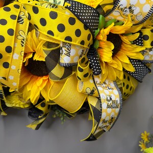 Peut inclure: Gros plan sur une couronne d&eacute;corative avec des tournesols, des rubans &agrave; pois jaunes et noirs et des rubans &agrave; carreaux. Les rubans portent les inscriptions "Bee Blessed" et "Cute as can bee". La couronne a un th&egrave;me d'abeille.