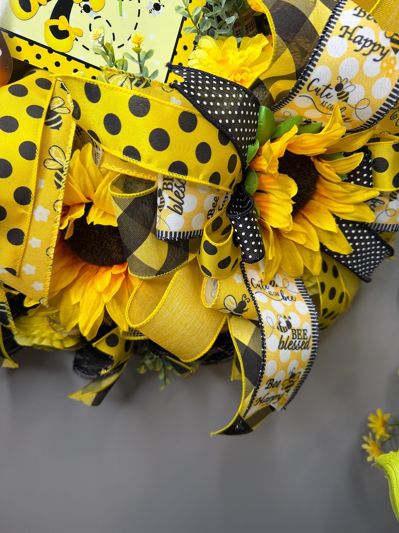 Peut inclure: Gros plan sur une couronne d&eacute;corative avec des tournesols, des rubans &agrave; pois jaunes et noirs et des rubans &agrave; carreaux. Les rubans portent les inscriptions "Bee Blessed" et "Cute as can bee". La couronne a un th&egrave;me d'abeille.