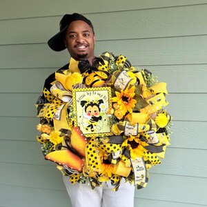Peut inclure: Une couronne d&eacute;corative avec des tournesols jaunes, des rubans et un panneau avec une abeille de dessin anim&eacute;. La couronne pr&eacute;sente des motifs &agrave; pois et &agrave; carreaux. Le panneau indique "Buzzin' by to say hi!"