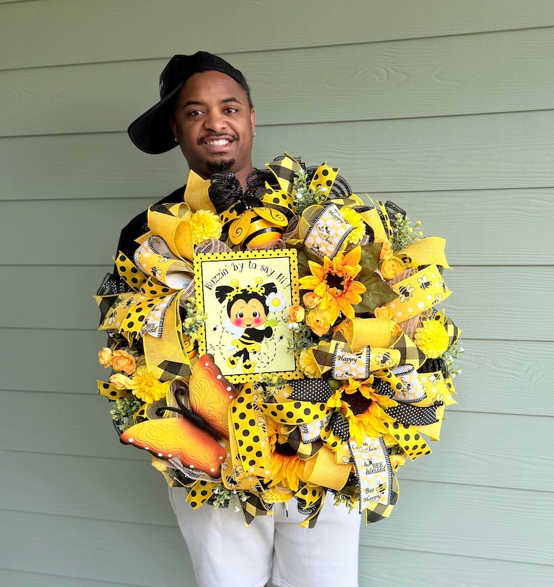 Peut inclure: Une couronne d&eacute;corative avec des tournesols jaunes, des rubans et un panneau avec une abeille de dessin anim&eacute;. La couronne pr&eacute;sente des motifs &agrave; pois et &agrave; carreaux. Le panneau indique "Buzzin' by to say hi!"