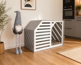 May include: A gray wooden pet crate with a slanted roof and white horizontal slats on the front panel. The crate features an open side design with diagonal white slats and contains a soft, dark-colored cushion inside for comfort.