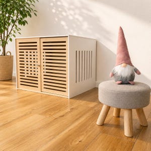 Könnte beinhalten: Eine Haustierbox aus weißem und hellem Holz mit horizontalen Lamellentüren und einem kleinen Metallriegel. Daneben steht ein runder grauer Stoffhocker mit hellen Holzbeinen, auf dem eine Plüsch-Gnom-Figur mit einem hohen, altrosa Filzhut sitzt.