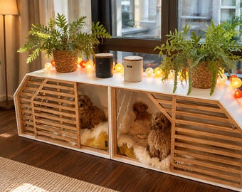 Op de afbeelding: Een wit en lichthouten huisdierconsole met twee compartimenten, elk voorzien van latten-schuifdeuren. Binnenin rusten twee kleine, krulharige honden op witte imitatiebontkussens. Het bovenoppervlak bevat twee cilindrische bussen en twee varens.