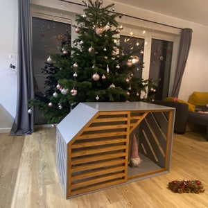 May include: A grey wooden dog house with a slatted wooden door and a gnome inside. The dog house is in front of a Christmas tree decorated with pink and silver baubles.