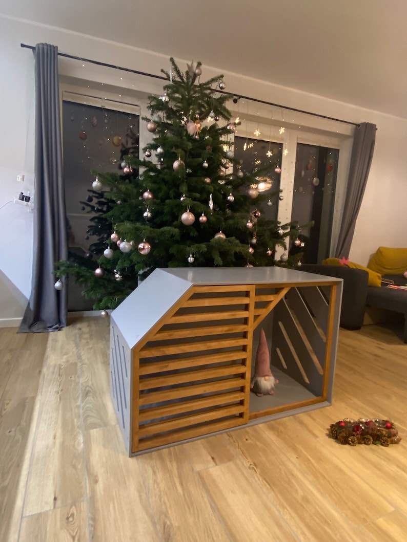 May include: A grey wooden dog house with a slatted wooden door and a gnome inside. The dog house is in front of a Christmas tree decorated with pink and silver baubles.