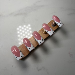 May include: A set of five press-on nails featuring a pink base with white tips and black crescent moon accents. The nails are arranged in a row on a wooden display stand, showcasing a glossy finish and a classic square-oval shape design.