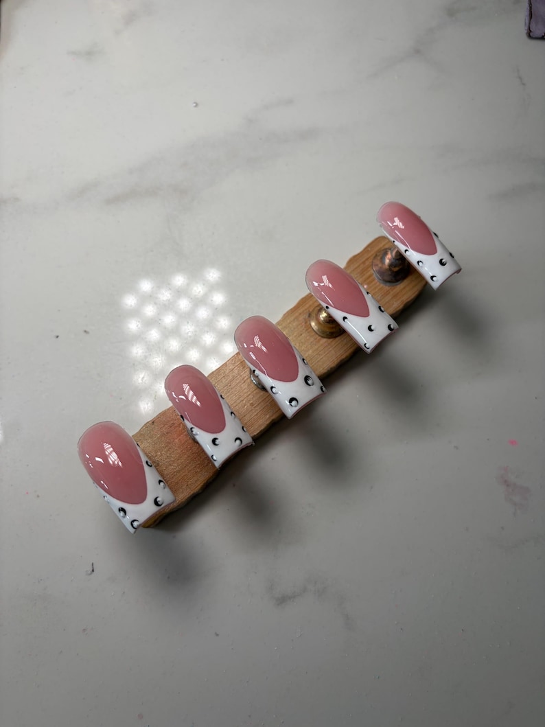 May include: A set of five press-on nails featuring a pink base with white tips and black crescent moon accents. The nails are arranged in a row on a wooden display stand, showcasing a glossy finish and a classic square-oval shape design.