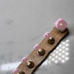 May include: A wooden display board with five artificial nails. Each nail has a light pink base with a white design on the tip. The nails are attached to small, round, gold-colored holders.