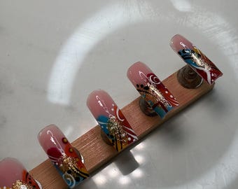 May include: Four press-on nails featuring a gradient base transitioning from clear to red. The design includes abstract swirls in blue, white, and black, accented with gold glitter flakes. The nails are displayed on a rectangular wooden stand.