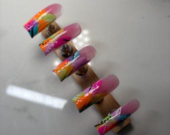 May include: Five press-on nails arranged on a wooden stand, each featuring a glossy finish with a vibrant abstract design. The nails display a mix of bright pink, orange, lime green, and blue paint strokes, accented with fine black lines and white dots.