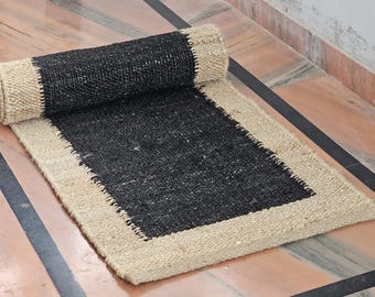May include: A rectangular jute rug with a black center and a natural beige border. The rug is partially rolled up, showcasing its texture and design. The rug is placed on a marble floor.