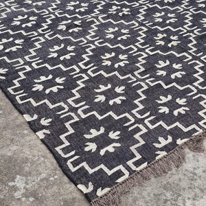 May include: A dark gray rug with a geometric pattern of white zigzags and floral-like shapes. The rug has a fringed edge and is placed on a concrete surface. This rug is suitable for home decor.