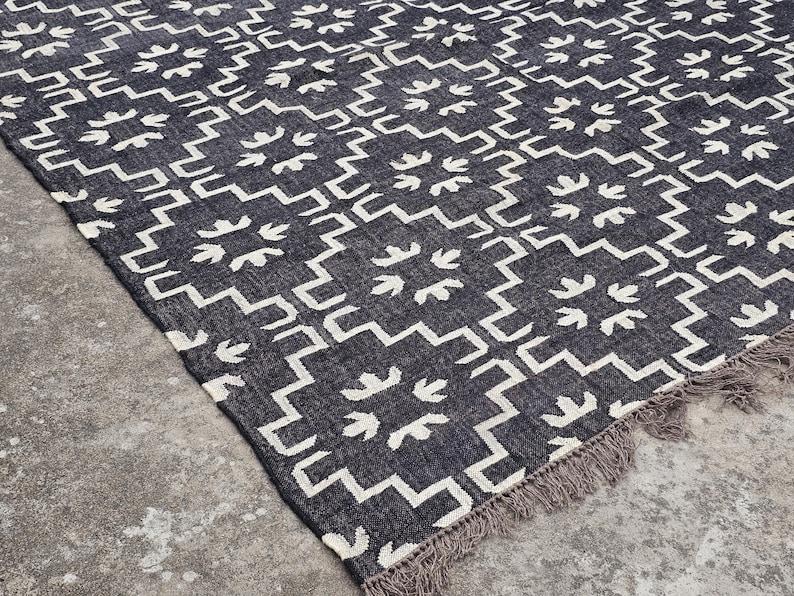 May include: A dark gray rug with a geometric pattern of white zigzags and floral-like shapes. The rug has a fringed edge and is placed on a concrete surface. This rug is suitable for home decor.