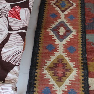 May include: A colorful rug with a geometric pattern in shades of red, blue, brown, and white. The rug has a black border and fringe on the ends.