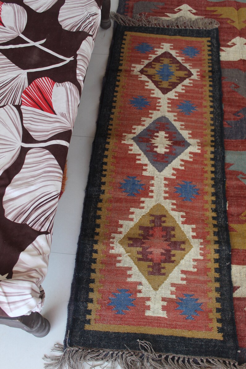 May include: A colorful rug with a geometric pattern in shades of red, blue, brown, and white. The rug has a black border and fringe on the ends.