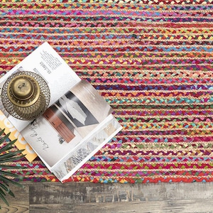 May include: A colorful braided rug with horizontal stripes in shades of red, pink, yellow, and blue. A magazine, a decorative brass lantern, and a potted plant are on the rug.