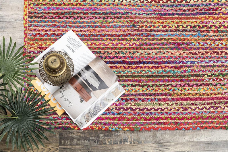 May include: A colorful braided rug with horizontal stripes in shades of red, pink, yellow, and blue. A magazine, a decorative brass lantern, and a potted plant are on the rug.