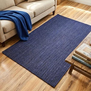 May include: A rectangular navy blue rug with a textured surface. The rug is placed on a hardwood floor, near a beige sofa and a wooden side table with books and a mug. A blue blanket is draped over the sofa.