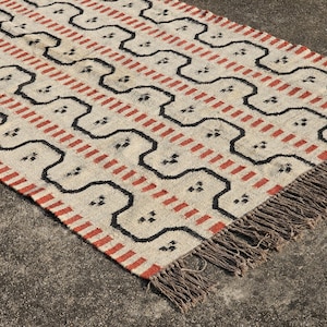 May include: Cream-colored rug with a repeating pattern of black and red geometric shapes. The rug has a fringed edge and is made of a woven material. The design features a series of connected shapes and stripes.
